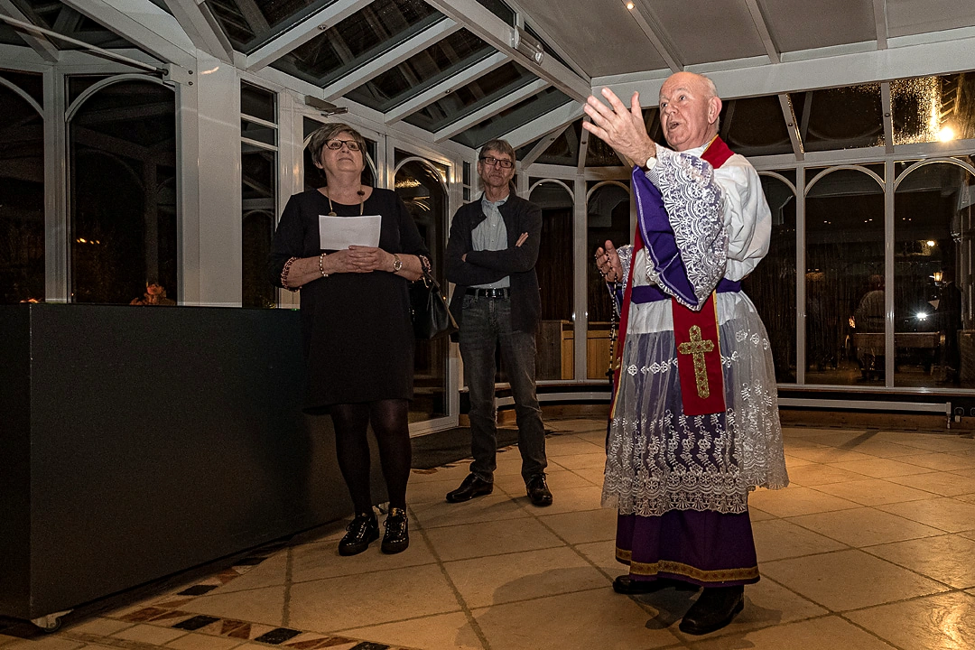 Priester in paarse en witte liturgische kledij spreekt in de glazen eventruimte